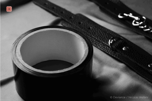 Black and white close-up of a roll of black bondage tape, with leather cuffs behind it