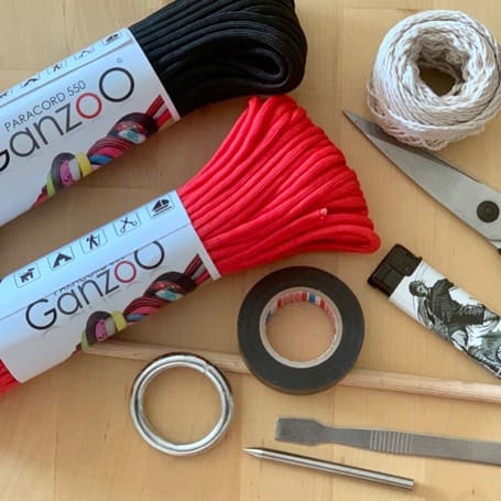 Materials for the DIY flogger on a wooden table: black and red paracord, a metal ring, insulating tape, twine, scissors, a lighter, a wooden dowel and tools