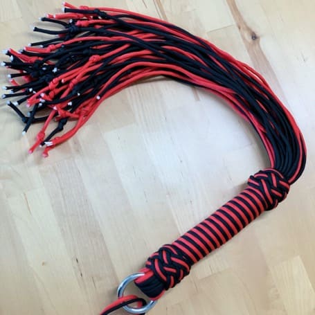 The finished DIY paracord flogger in black and red, with a braided handle and metal ring, on a wooden table