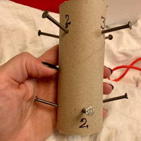 A hand holding a cardboard tube with numbered nails pushed through it as a weaving jig