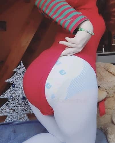 Person in a red top with striped sleeves wearing a patterned nappy, beside a teddy bear and a silver Christmas tree