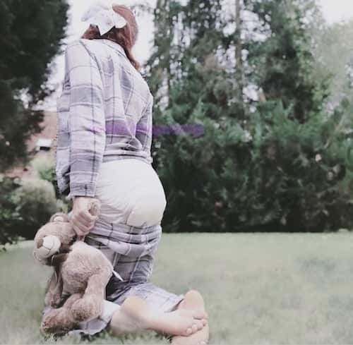 Person in a checked shirt kneeling barefoot in a garden, wearing a nappy and holding a plush rabbit