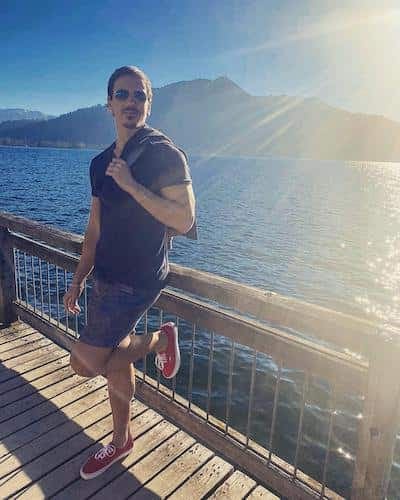 Person in sunglasses and a dark shirt standing backlit on a wooden jetty by a mountain lake