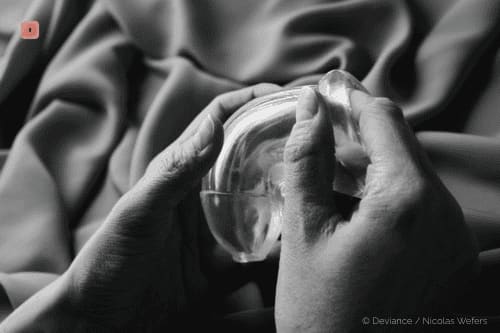 Black and white close-up of two hands holding a clear silicone cock ring against folded fabric