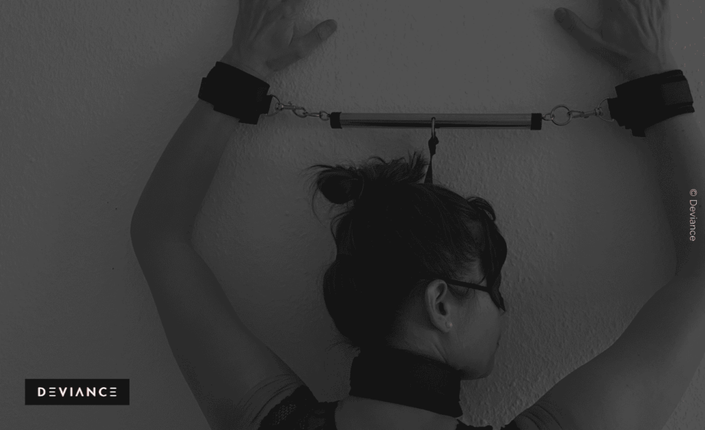 A person with bound hands standing against a wall, while the small spreader bar holds their arms in the desired position.